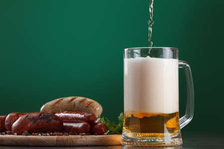 Traditional Oktoberfest menu. Beer being poured into a glass with foam, traditional snacks, sausages grilled. Copyspace with green backgroundの写真素材
