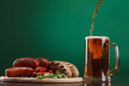 Traditional Oktoberfest menu. Beer being poured into a glass with foam, traditional snacks, sausages grilled. Copyspace with green backgroundの写真素材