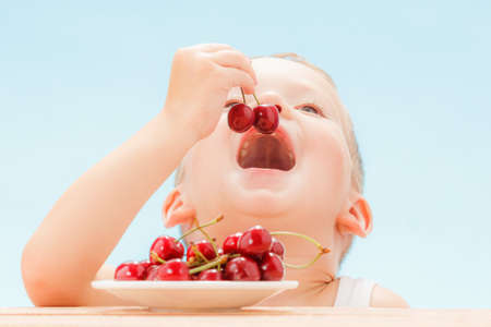 Child with red berries, boy eats and chooses cherry. Blue sky background.の写真素材