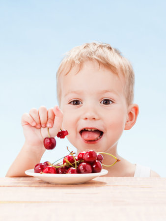 Child with red berries, boy eats and chooses cherry. Blue sky background.の写真素材