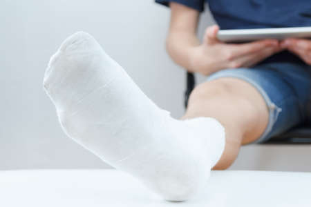 Man with broken leg in a plaster sits on chair and reads the tablet, white backgroundの写真素材