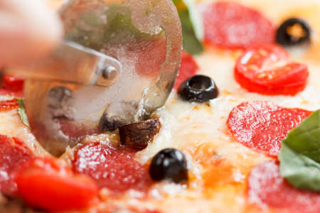 Cutting pepperoni pizza on slices with special knife. Fresh salad at background. Hand holding knife.の写真素材