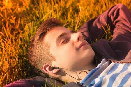 Portrait of a cute teen boy listening to music, lying down on a fresh green grass field, happy summer holidaysの写真素材