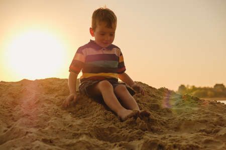 Four-year-old caucasian boy playing in the sand on the beach, summer sunset on the river bank in the sunlight sunbeamの写真素材