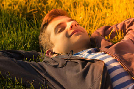 Portrait of a cute teen boy listening to music, lying down on a fresh green grass field, happy summer holidaysの写真素材