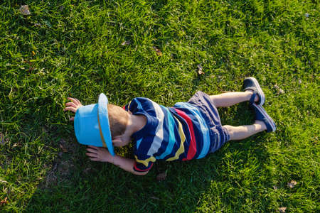 Handsome little boy lying on grass. Little dreamer lies in meadow with hat. Child in nature outside city. Carefree pastime, vacationの写真素材