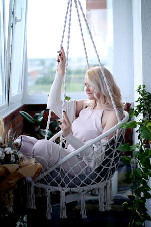 Beautiful blonde sits on a balcony in a boho style armchair among greeneryの写真素材