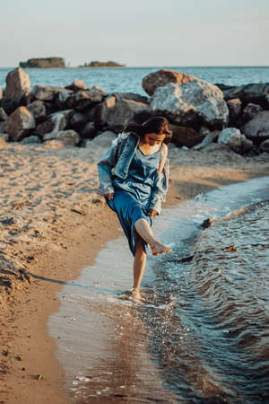 A girl in a blue dress walks along the coastline of the beach. Summer at sea, walk at sunset.の写真素材