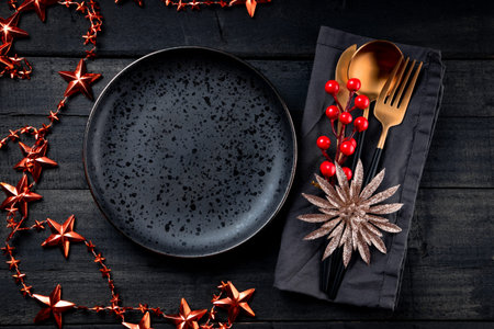 Christmas background - golden cutlery on a linen napkin and a black empty plate on a black wooden...