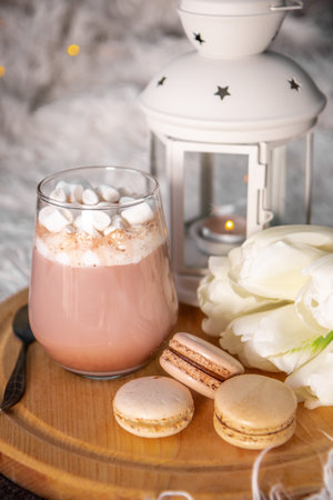 A glass of hot cocoa with marshmallows, macaroni cakes and white tulips on a tray in bed close up,...
