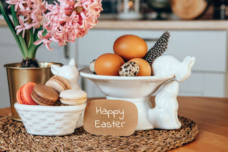 Postcard with the inscription Happy easter. Easter eggs, macaroon cakes, pink hyacinth and porcelain Easter bunnies on the table in the interior kitchenの写真素材
