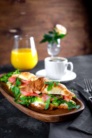 Croissants with arugula, avocado and salmon, a cup of black coffee and a glass of orange juice on the table, breakfast concept with soft focus vertical photoの写真素材