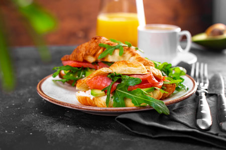 Croissants with arugula, avocado and salmon, a cup of black coffee and a glass of orange juice on the table, breakfast concept with soft focusの写真素材