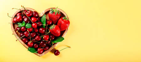 Ripe cherries and strawberries on a wooden plate in the shape of a heart on a yellow background, web banner with free space for textの写真素材