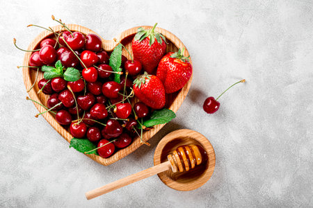 Ripe cherries and strawberries on a wooden plate in the shape of a heart and honey on a light gray background. High quality photoの写真素材
