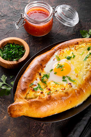 A traditional dish of Georgian cuisine, Adjarian khachapuri with cheese and egg and spicy tomato sauce adjika on the dark background close up vertical photoの写真素材