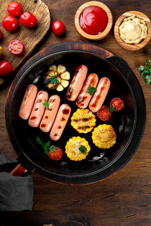 Grilled sausages with vegetables and sauces. Summer BBQ with sausages, corn and tomatoes on the wooden table top view vertical photoの写真素材