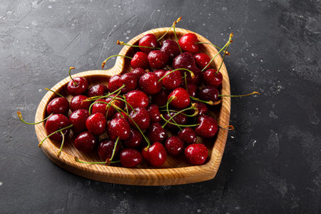 Ripe cherries and strawberries on a wooden plate in the shape of a heart and dark gray background,...