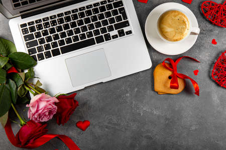 A laptop, cup of coffee, cake in shape of heart and bouquet of red and pink roses on the desk top view. Valentines card with copy space for textの写真素材