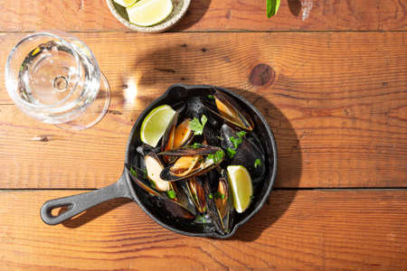 Black mussels in garlic sauce with herbs and lime and a glass of white wine on the wooden table under the summer sun top viewの写真素材