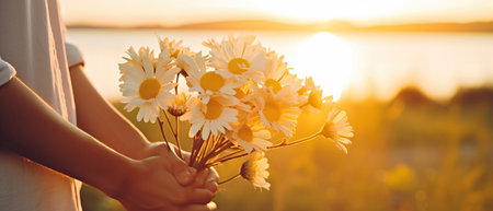 A person is holding a beautiful bouquet of daisies, showcasing the delicate petals and vibrant colors against a backdrop of a natural landscape with green grass and a clear blue skyの素材