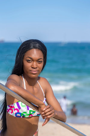 Young black woman at the beach, wearing a bikini. Leaning on railing. Blue sky.の写真素材