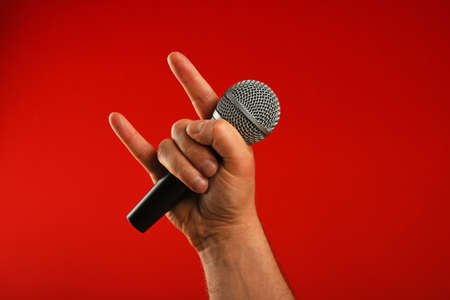 Man hand holding voice microphone with devil horns rock metal gesture sign over red background, diagonalの写真素材