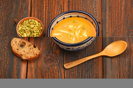 Ceramic bowl of pumpkin cream soup, spoon, slice of bread and seeds on dark wooden table background,...