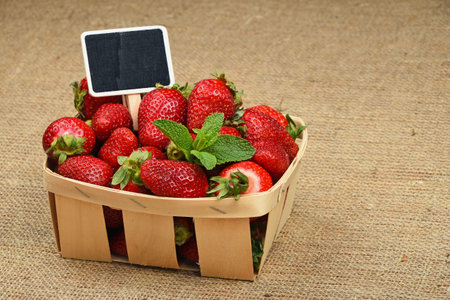 Wicker wooden basket of strawberries with mint leaves and chalk blackboard price tag sign on jute burlap canvas background, high angle viewの写真素材
