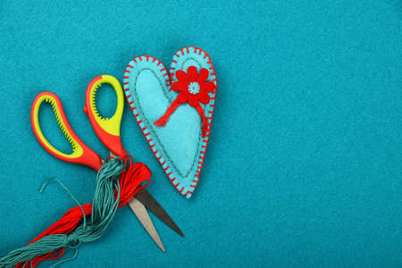 Craft and art, one blue handmade stitched toy heart, red thread and scissors on teal backgroundの写真素材