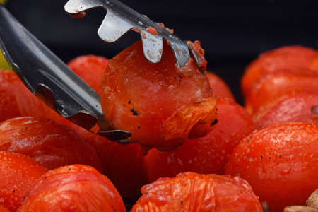 BBQ small red tomatoes cooked on barbecue, tongs holding one tomato, picking it up from grillの写真素材