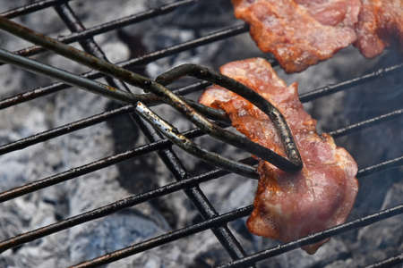 Tongs holding crispy smoked grilled barbecue bacon slice, cooked on bbq smoke grill, close upの写真素材