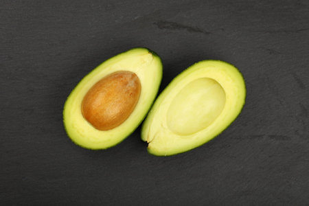 One fresh ripe green avocado cut in two halves with pit stone on black slate board background,...