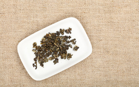 White small plate with dried green tea leaves on flax canvas tablecloth, close up, elevated top...