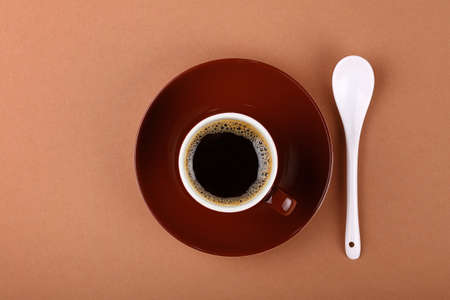 Full small brown ceramic espresso cup of black coffee with saucer and white spoon on paper parchment, close up, elevated top view, directly aboveの写真素材