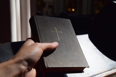 Man hand holding Holy Bible old vintage book in canvas hardcover over wooden pew during prayer in Christian catholic church or cathedral, close up, personal perspectiveの写真素材