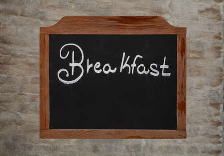 White chalk written word BREAKFAST over old black school chalkboard blackboard sign in vintage brown wooden frame over background of grunge uneven gray daub plaster wall, close upの写真素材