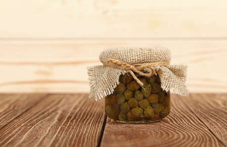 Close up of one small glass jar of pickled capers with canvas top decoration and twine on brown wooden table over white background, low angle side viewの写真素材