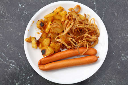Close up portion of two boiled sausages with roasted potato and fried onion rings on white plate over grey table, elevated top view, directly aboveの写真素材