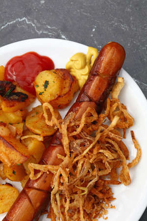 Close up portion of one big grilled sausage with homemade roasted potato, fried onion rings, ketchup and mustard on white plate over grey table, elevated top view, directly aboveの写真素材