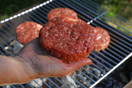 Close up hand holding raw beef or pork meat barbecue burgers for hamburger to cook grilled on bbq grill, high angle viewの写真素材
