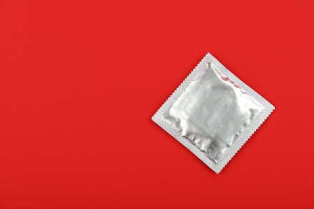 Close up one condom silver wrap pack over vivid red background with copy space, elevated high angle...