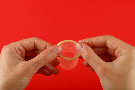 Close up two woman hands holding one open latex condom over red background with copy space, low...