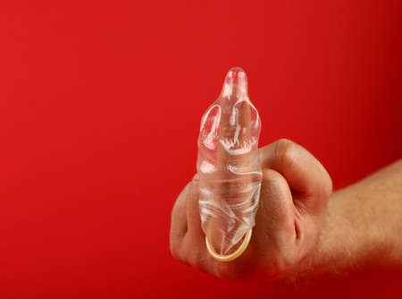 Close up man hand showing protest or insult rude gesture with latex condom on finger over red background with copy space, low angle side viewの写真素材