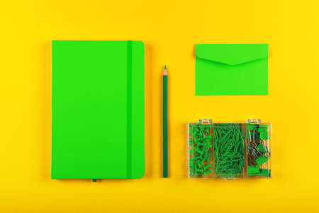 Neatly organized stationery flat lay of green notebook, envelope, pencil, office clips, binders and pins in box over yellowの写真素材