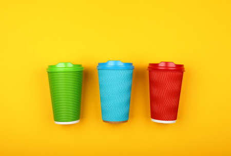 Close up three crimped disposable paper takeaway coffee cups, red, green and blue, with plastic cap over vivid yellow background, flat lay, elevated top view, directly aboveの写真素材
