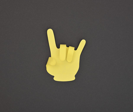 Paper made yellow DEVIL HORNS hand gesture sticker over grey background