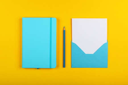 One open blank pastel blue envelope with white paper, closed notebook and wooden pencil over vivid yellow background, flat lay, directly aboveの写真素材