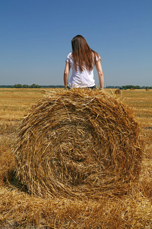 Young Caucasian woman sitting on the straw bale in stubble field after harvesting, low angle rear...