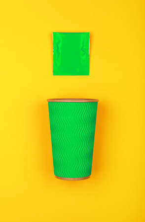 Close up one crimped disposable green paper takeaway cup and instant coffee, sugar or teabag sachet over vivid yellow background, flat lay, elevated top view, directly aboveの写真素材